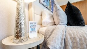 Ein Bett mit weißem Kopfteil, einer grauen Decke und einem schwarzen Kissen. Auf einem kleinen Tisch steht ein Schild mit der Aufschrift 'SHARE YOUR Stay in my District' und Social-Media-Symbolen.