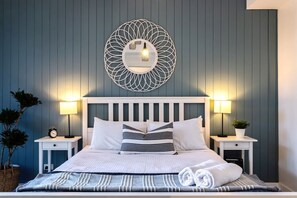 A neatly made bed with a striped blanket, two bedside lamps, and a wall-mounted mirror with a geometric design.
