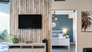 A living room with a wooden wall, a mounted TV, a white entertainment unit, and a doorway leading to a bedroom with a bed and a nightstand. - `Family Beach Getaway` by Inn Paradise (Surfers Paradise)