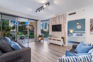 A modern living room with a large TV, a balcony with a view of buildings, and a dining area with outdoor furniture.