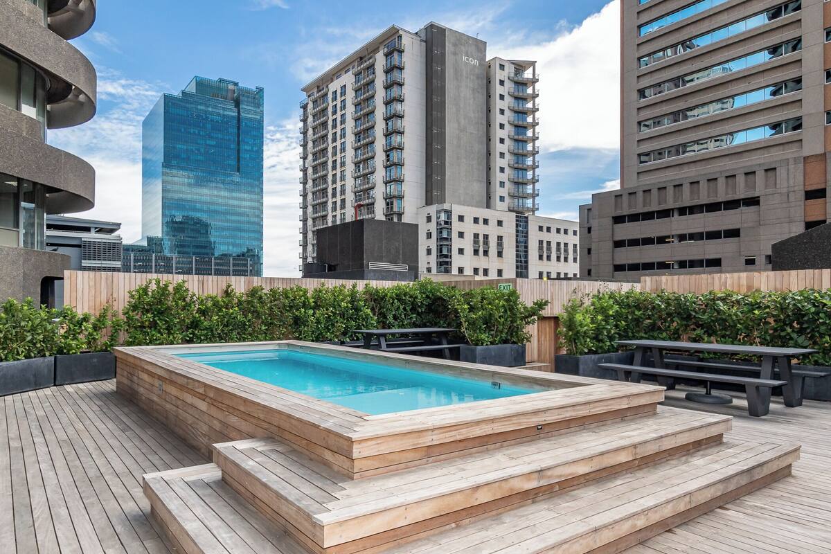 A rooftop pool with wooden steps, surrounded by greenery and modern buildings in the background.