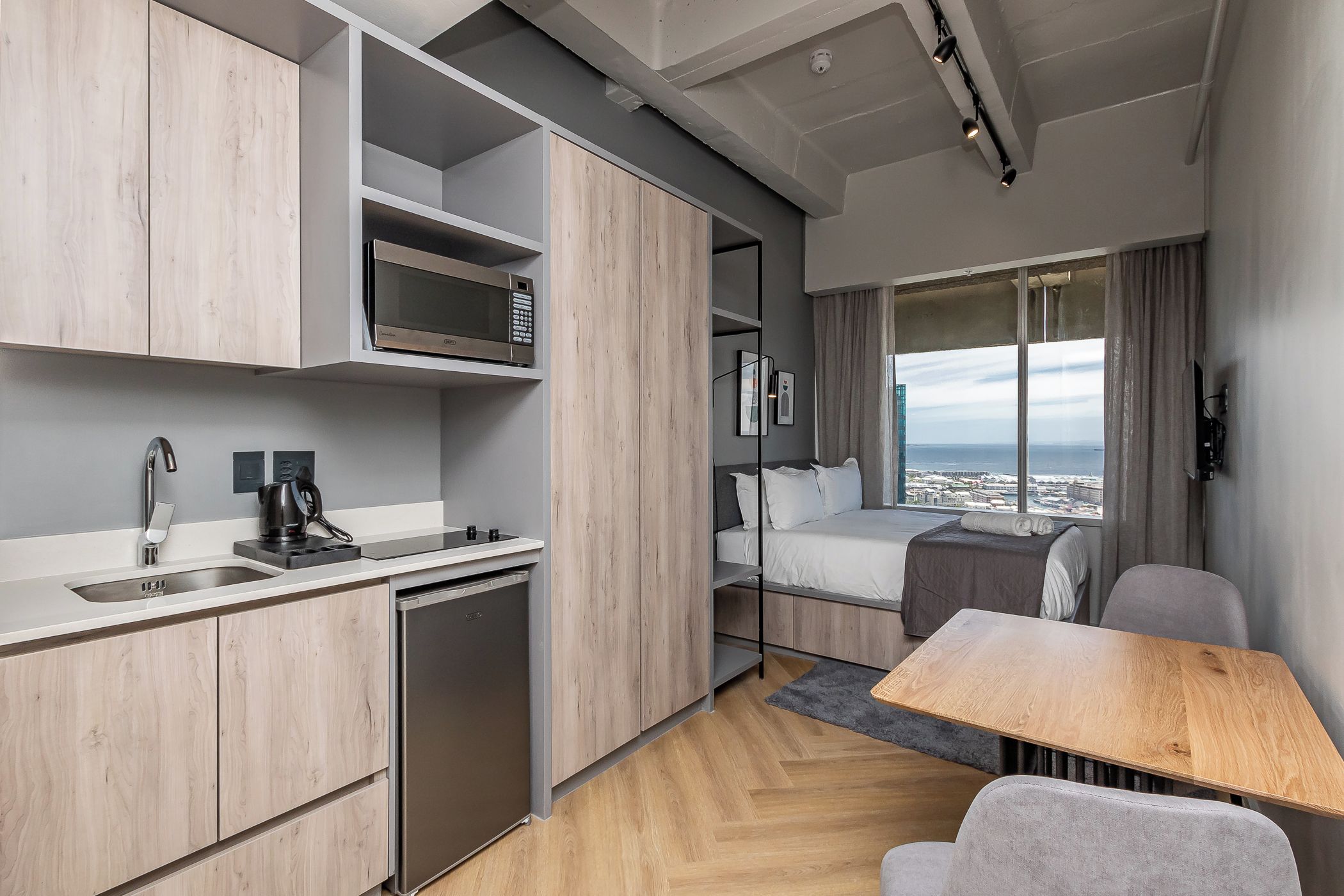 A compact hotel room with a kitchenette, a bed with a city view, and a small dining area.