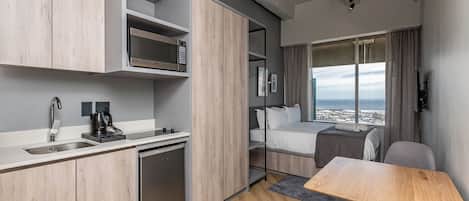 A compact hotel room with a kitchenette, a bed with a city view, and a small dining area.
