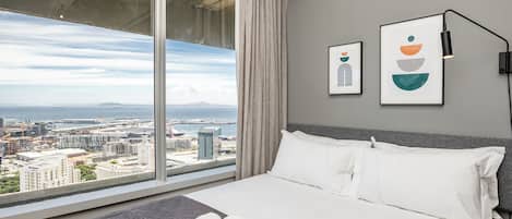A hotel room with a large window offering a city view, a neatly made bed with white linens, and two framed artworks on the wall.