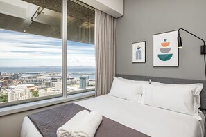 A hotel room with a large window offering a city view, a neatly made bed with white linens, and two framed artworks on the wall. - One Thibault Residence By Itc Hospitality (Cape Town)