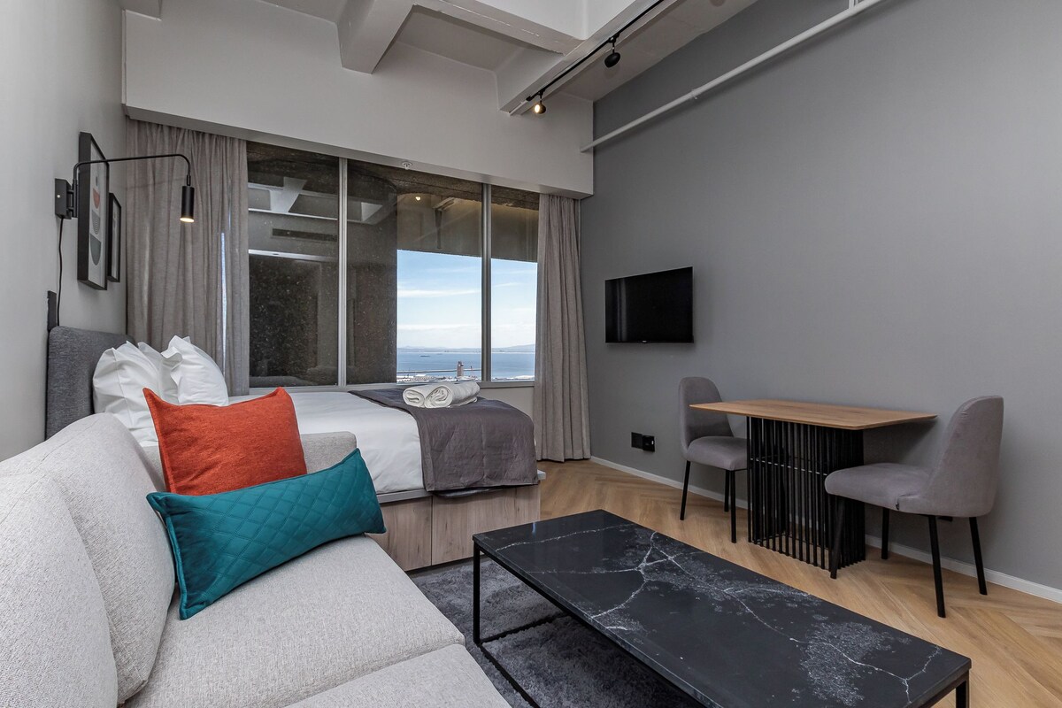 A modern hotel room with a large bed, a sofa, a dining table, and a TV mounted on the wall.