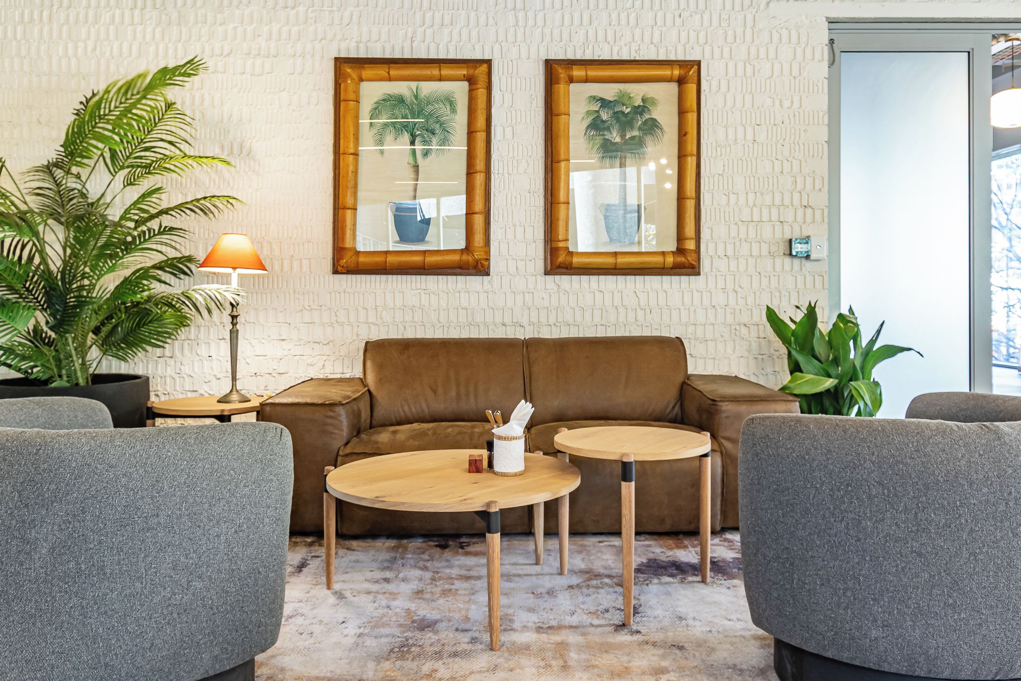A living room with a brown sofa, two round wooden tables, and two armchairs. There are two framed pictures of potted plants on the wall.