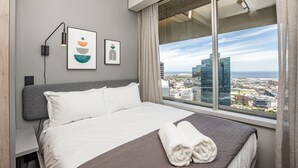 A modern hotel room with a large bed, a city view, and artwork on the wall.