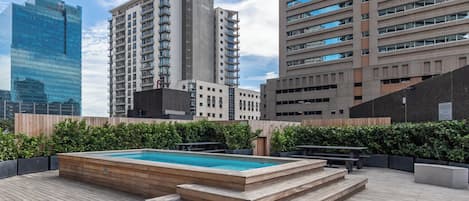 A rooftop pool area with wooden steps leading to the pool, surrounded by greenery and overlooking a cityscape with modern buildings.