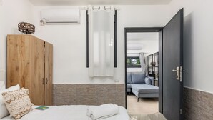 Ein Schlafzimmer mit Holzschrank, einem Bett mit weißer Bettwäsche, einem Badezimmer mit Duschvorhang und Blick in einen Wohnbereich mit einer Couch.