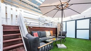 Eine Terrasse im Freien mit Holzterrasse, einem grauen Sofa mit Kissen, einem Glastisch mit einer Wasserflasche und Obst sowie einem großen Sonnenschirm.
