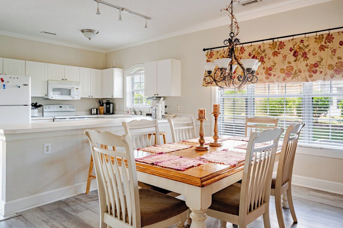 Una cocina con armarios blancos, una mesa de comedor de madera con sillas y una ventana con persianas.