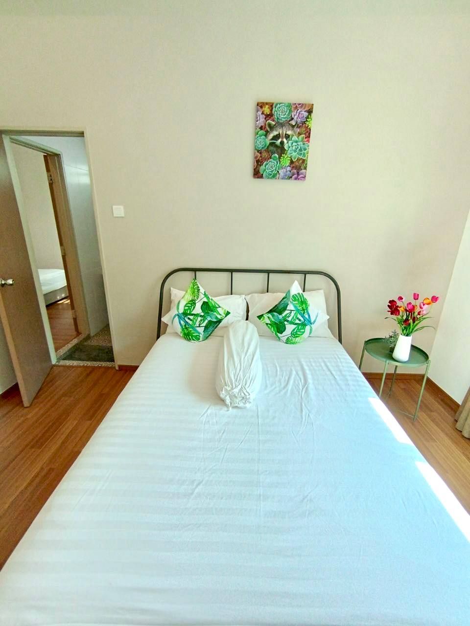 A neatly made bed with white linens and green patterned pillows, a small bedside table with a vase of flowers, and a painting on the wall.