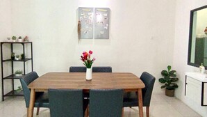A dining area with a wooden table and six chairs, a ceiling fan, a wall-mounted light, a small shelf with plants, and a potted plant near the window. - Tropical Livings Meru Ipoh Homestay (Ipoh)