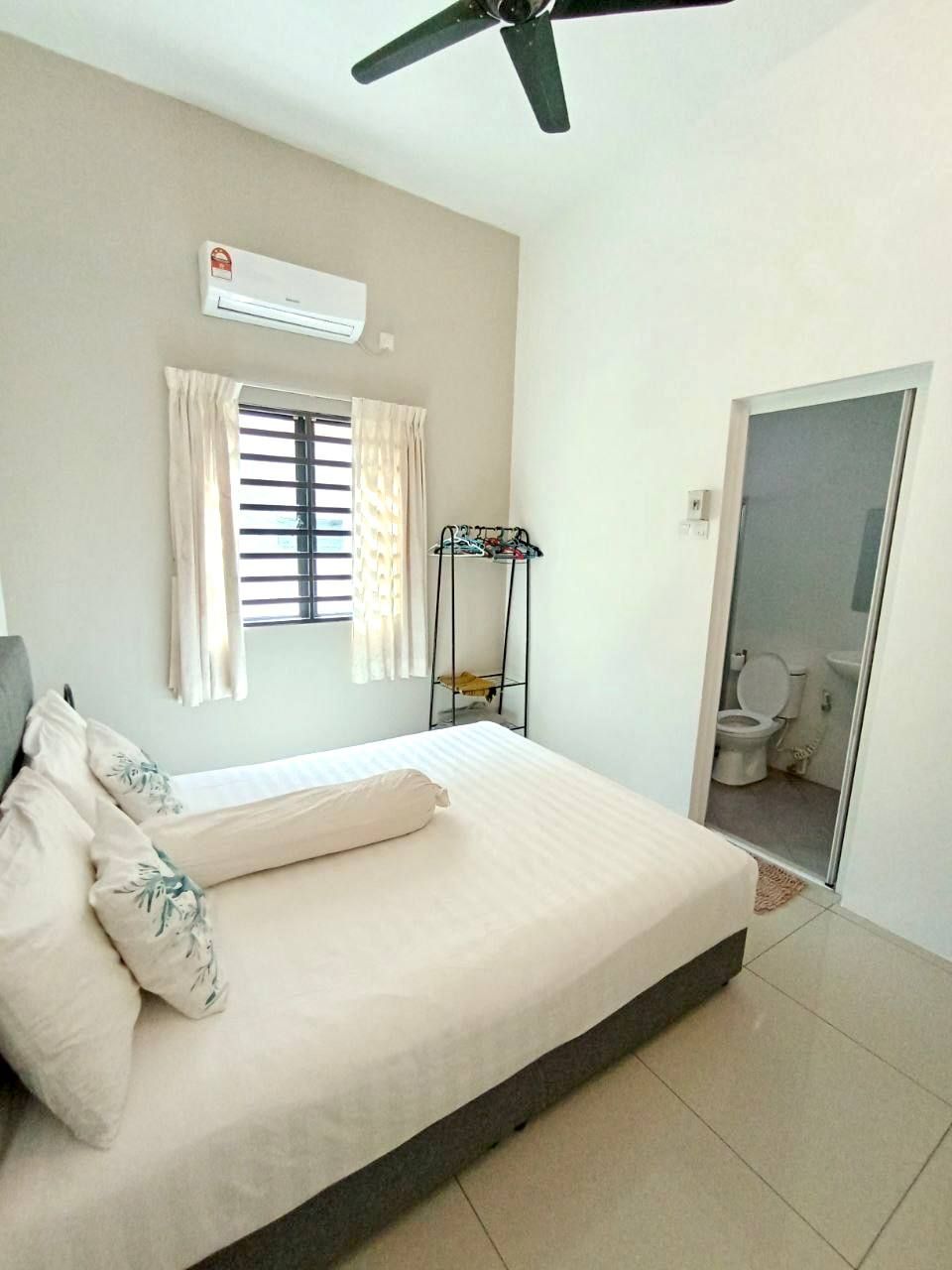 Un dormitorio con cama, aire acondicionado, ventana con cortinas, ventilador de techo y baño visible a través de una puerta abierta.
