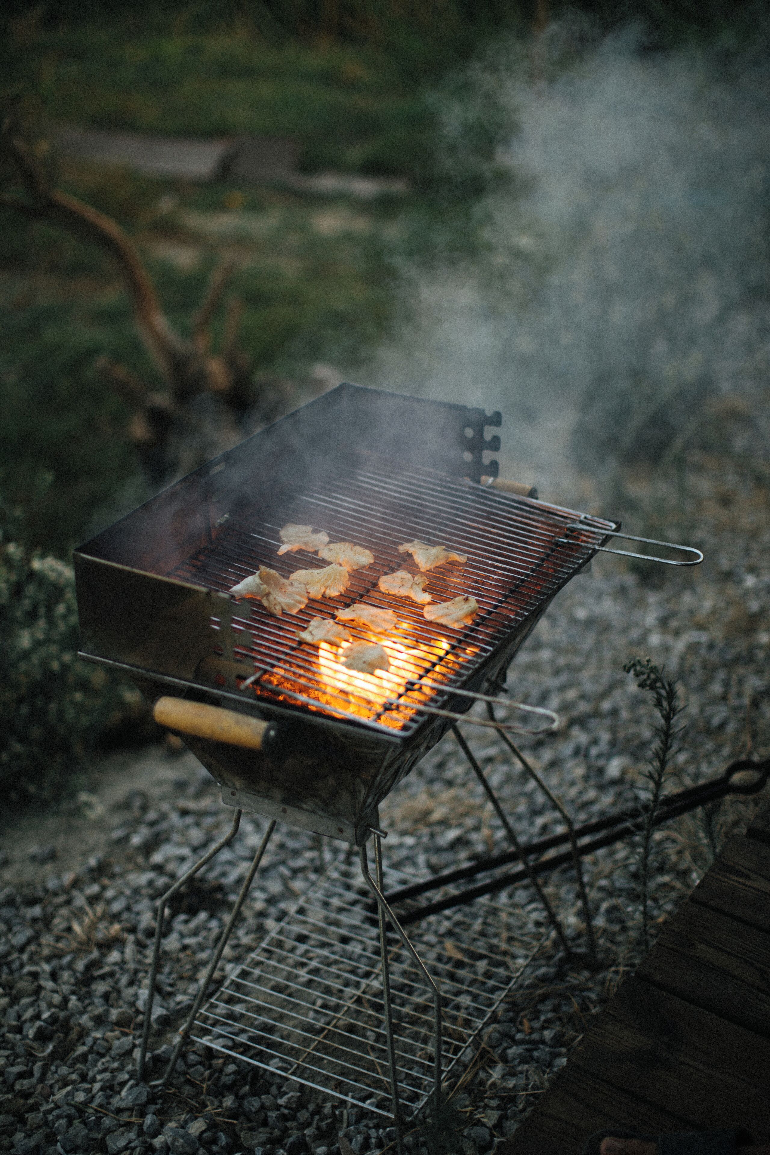 Una parrilla portátil con comida cocinándose, sobre una superficie de grava al aire libre.