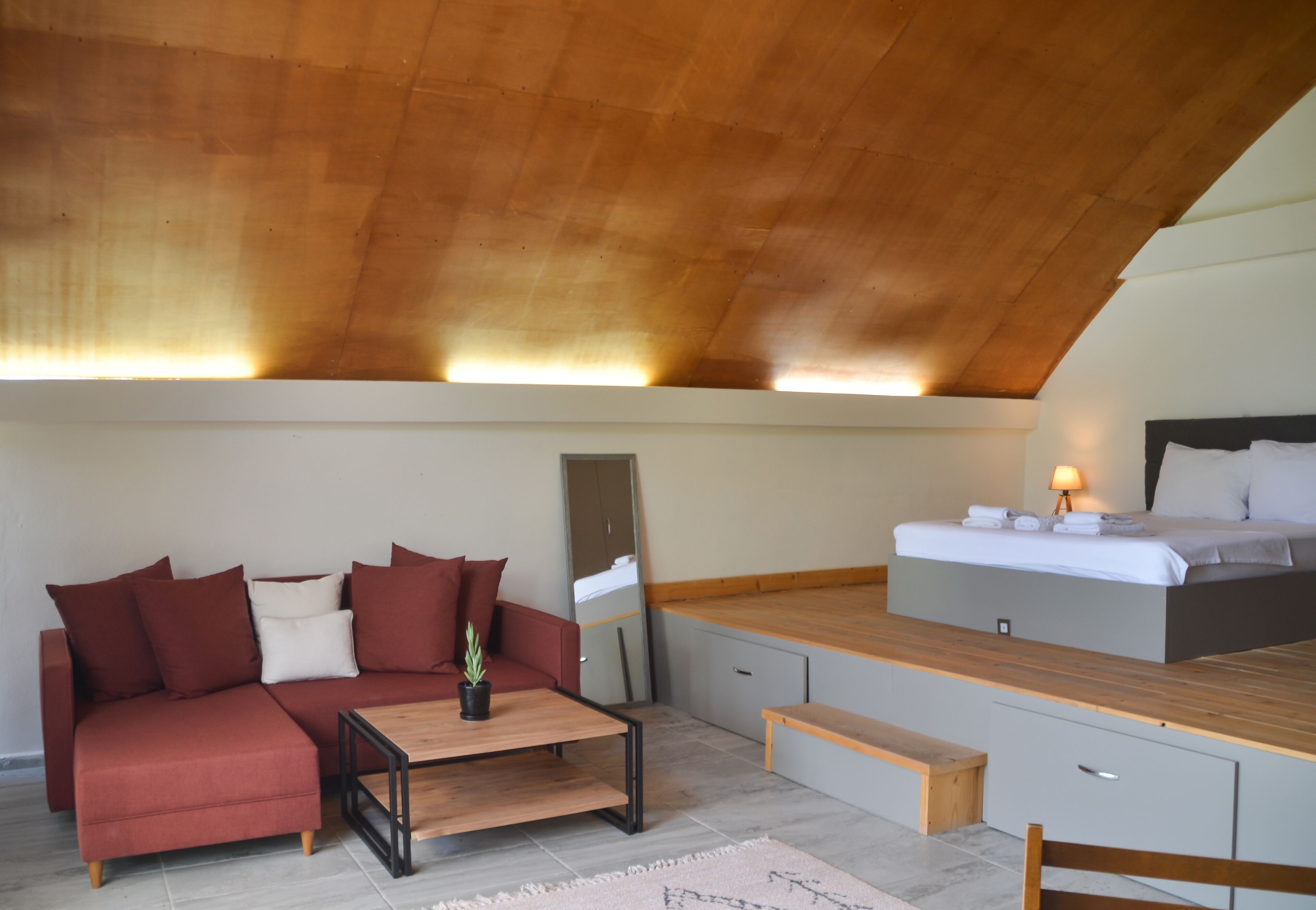 Un dormitorio tipo loft moderno con techo de madera, un sofá rojo, una mesa de centro de madera, una cama con mesita de noche y un escritorio pequeño.