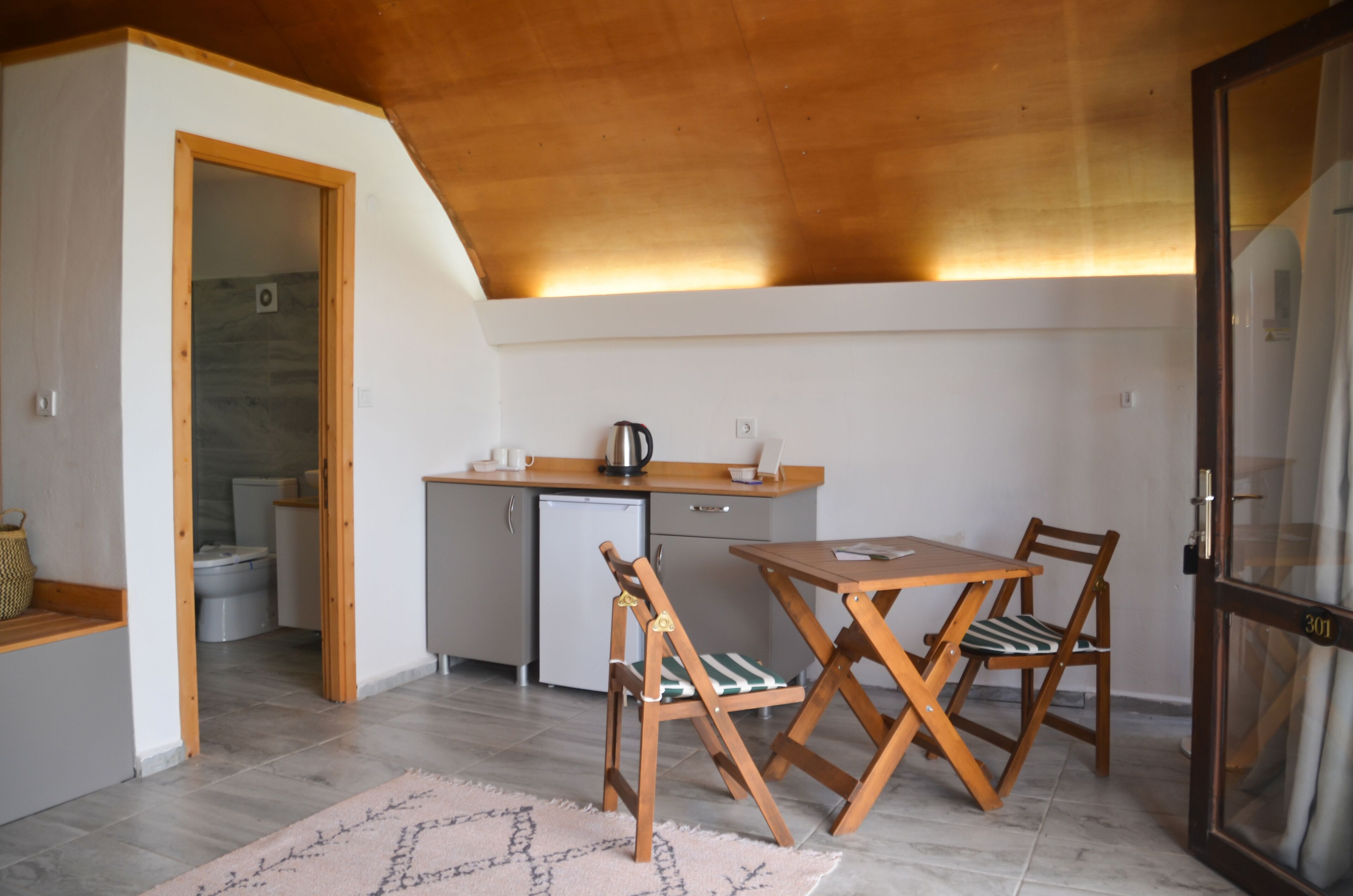A compact kitchen with a wooden table and chairs, a small refrigerator, and a coffee maker.