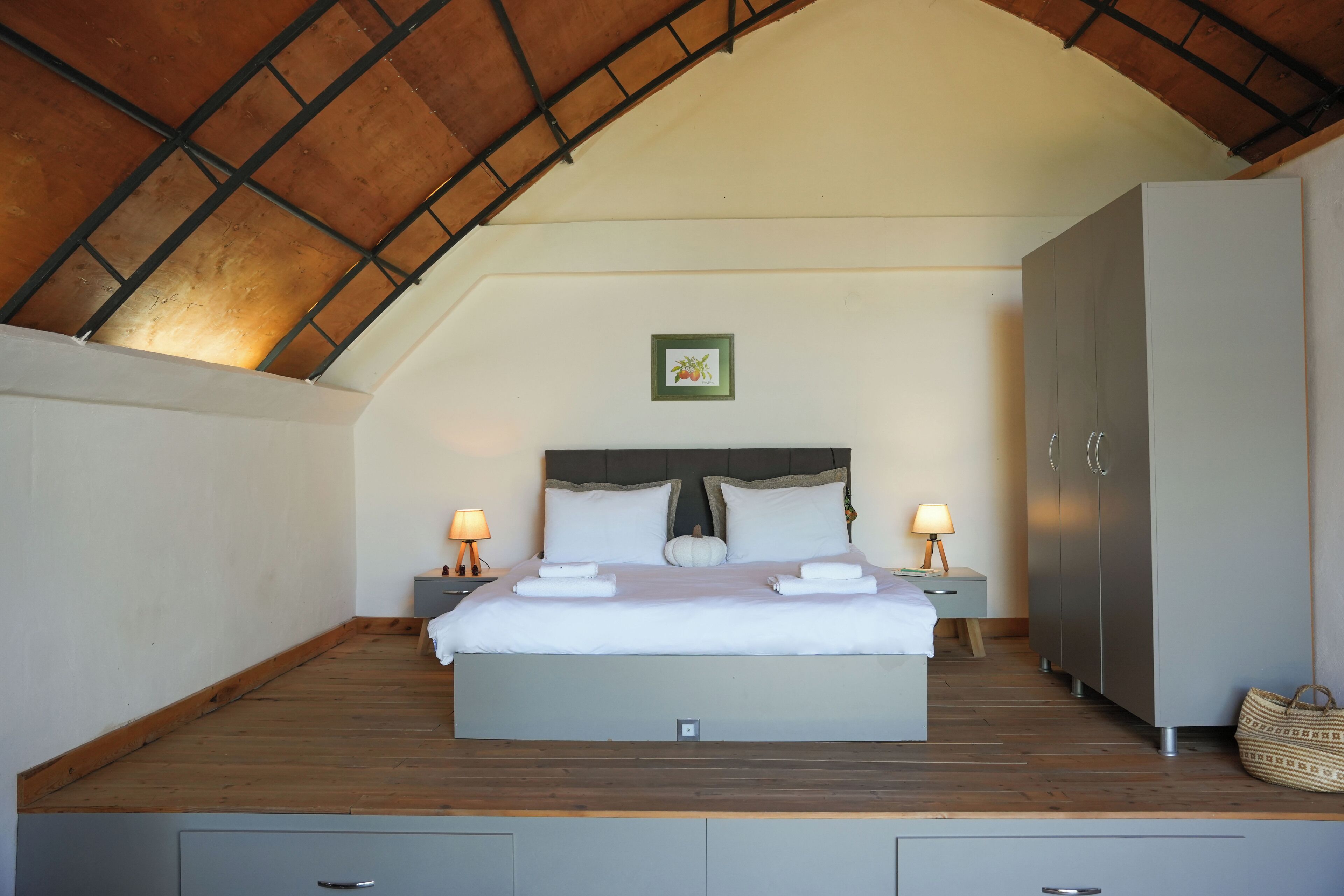 Een slaapkamer met een schuine kap, een bed met witte lakens, twee nachtkastjes met lampen, een garderobe en een klein lijstwerk met een afbeelding aan de muur.
