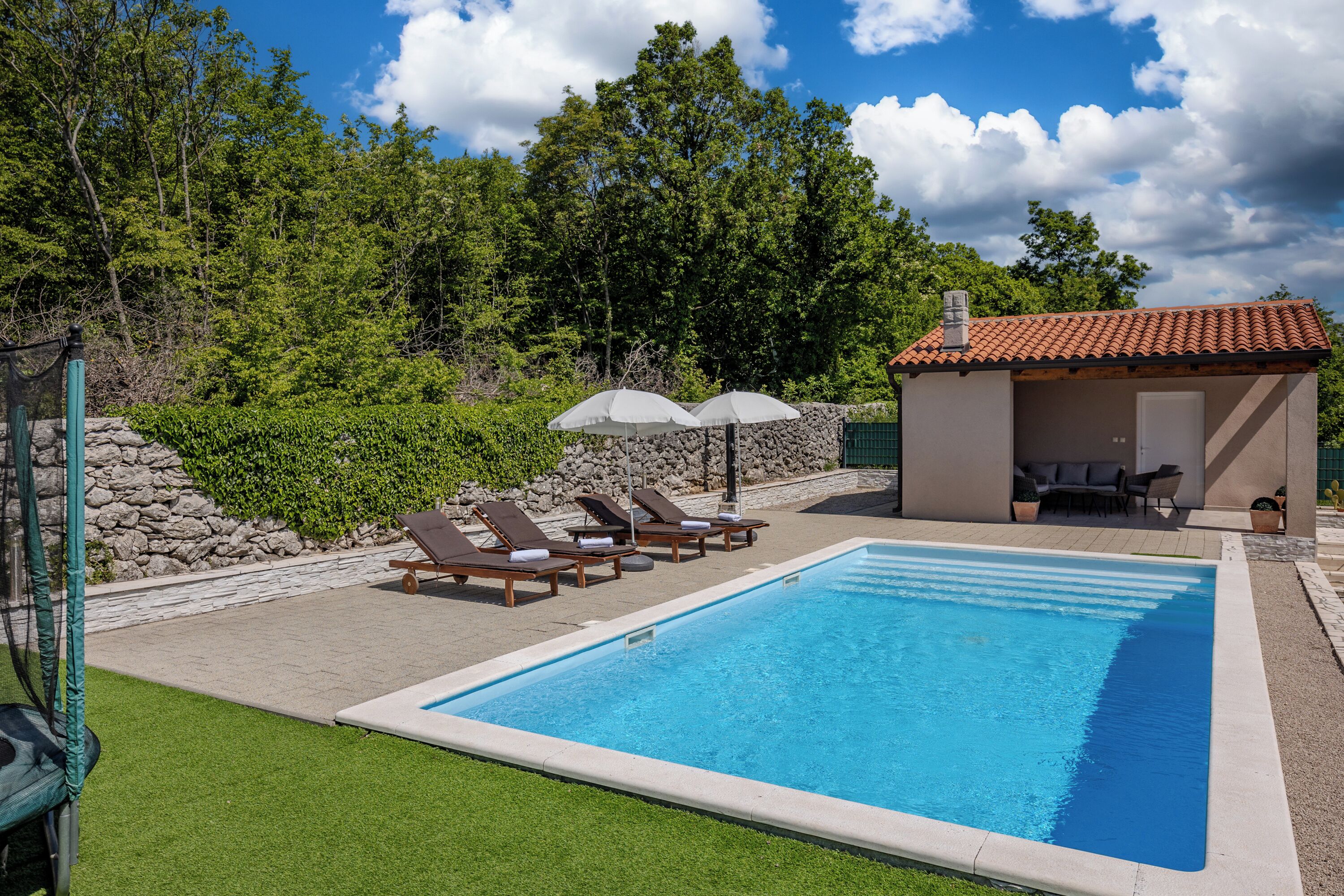 Une piscine rectangulaire, agrémentée de chaises longues et de parasols, entourée d’un mur de pierre et de verdure.