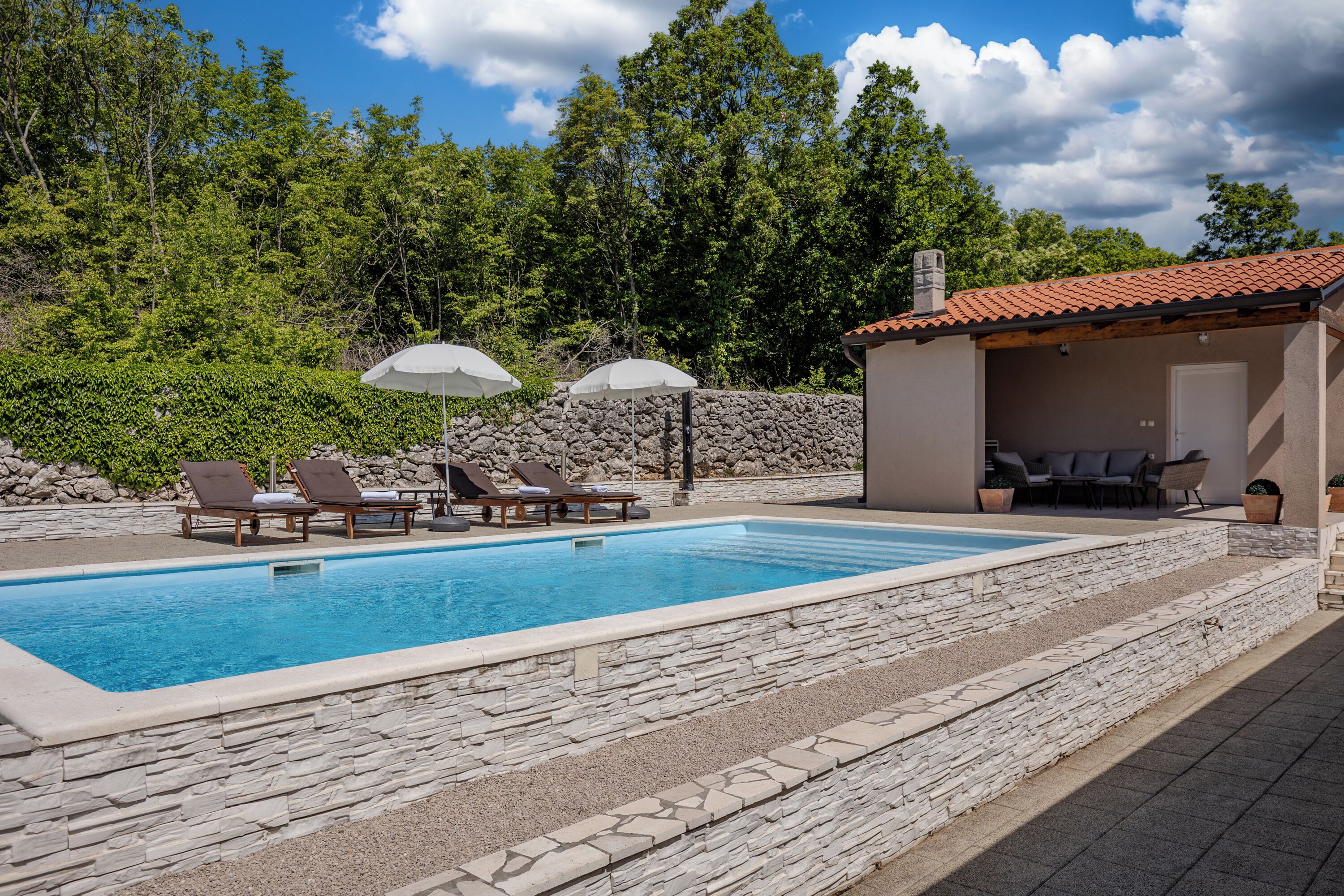 Una piscina con sillas de descanso y sombrillas, rodeada de muros de piedra y una escalera de piedra.