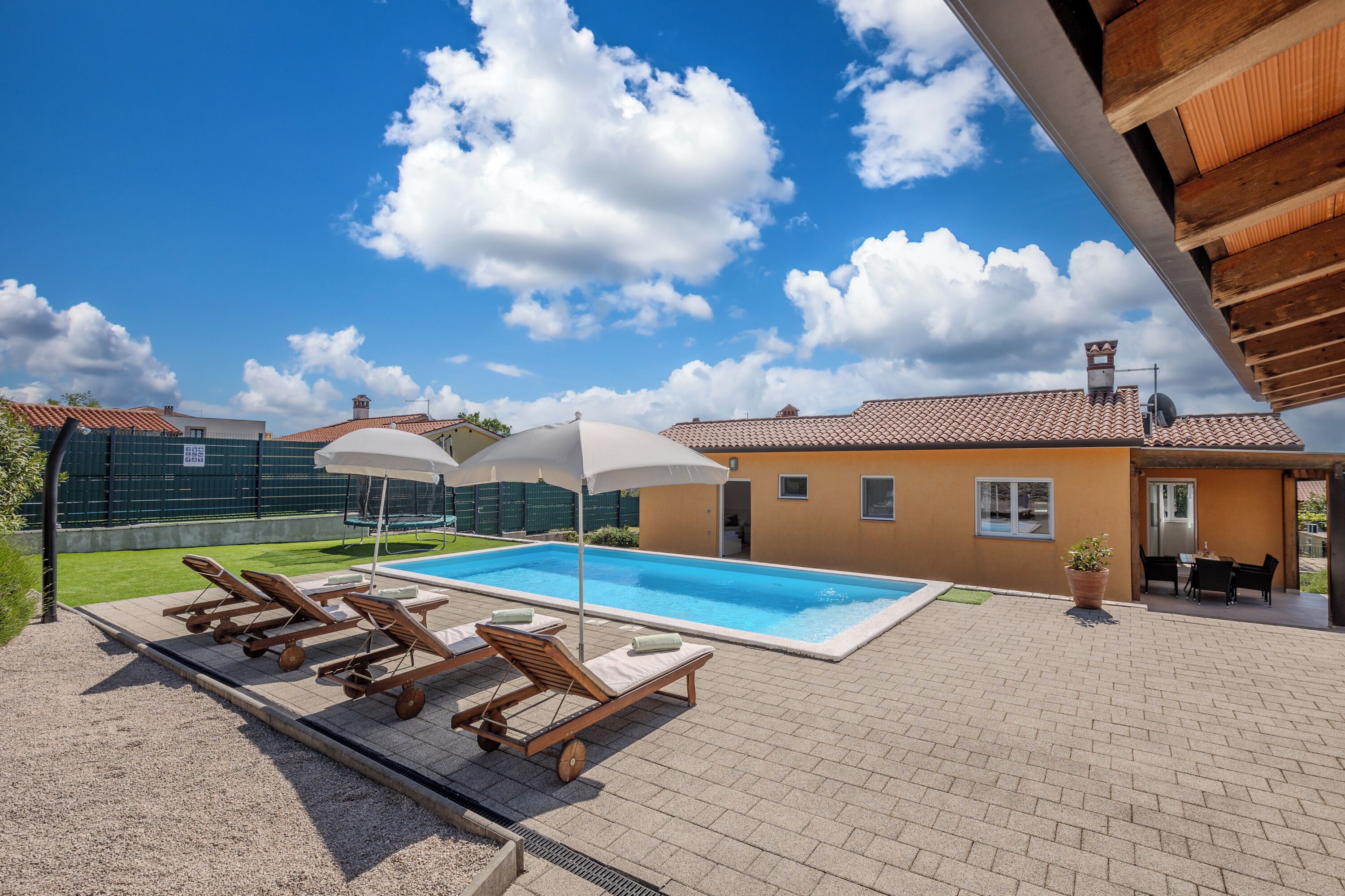 A swimming pool with lounge chairs and umbrellas, surrounded by a brick patio and a wooden pergola.
