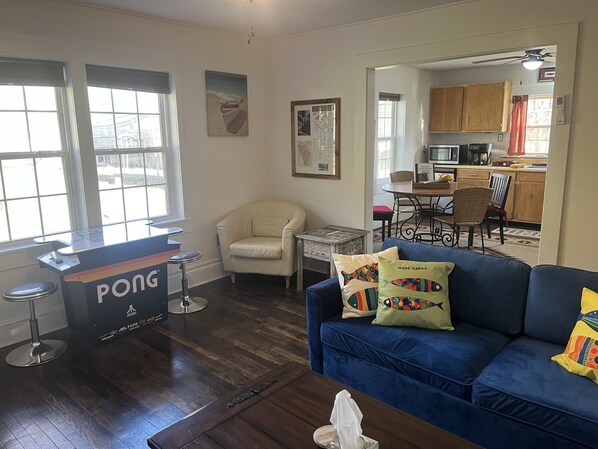 A living room with a blue sofa, a white armchair, a small coffee table, and a ping pong table. There is a kitchen visible through an open doorway.