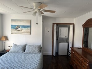 A bedroom with a bed, a ceiling fan, a painting of a boat on the wall, a dresser, and a door leading to a bathroom.