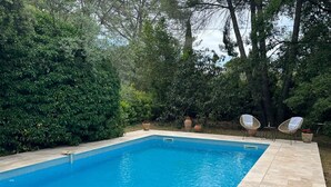 A rectangular swimming pool with a tiled edge, surrounded by a stone patio. There are two wicker chairs with white cushions and potted plants nearby. - Family House in Provence with Pool and Garden (Draguignan)