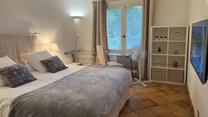 A hotel room with a bed, a desk, a chair, a standing fan, a TV, and a shelving unit with decorative items. - Family House in Provence with Pool and Garden (Draguignan)