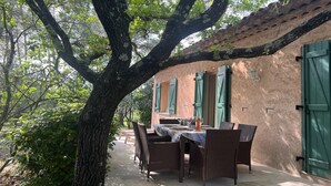 An outdoor dining area with wicker furniture and a table set for two, surrounded by trees and a rustic building with green shutters. - Family House in Provence with Pool and Garden (Draguignan)