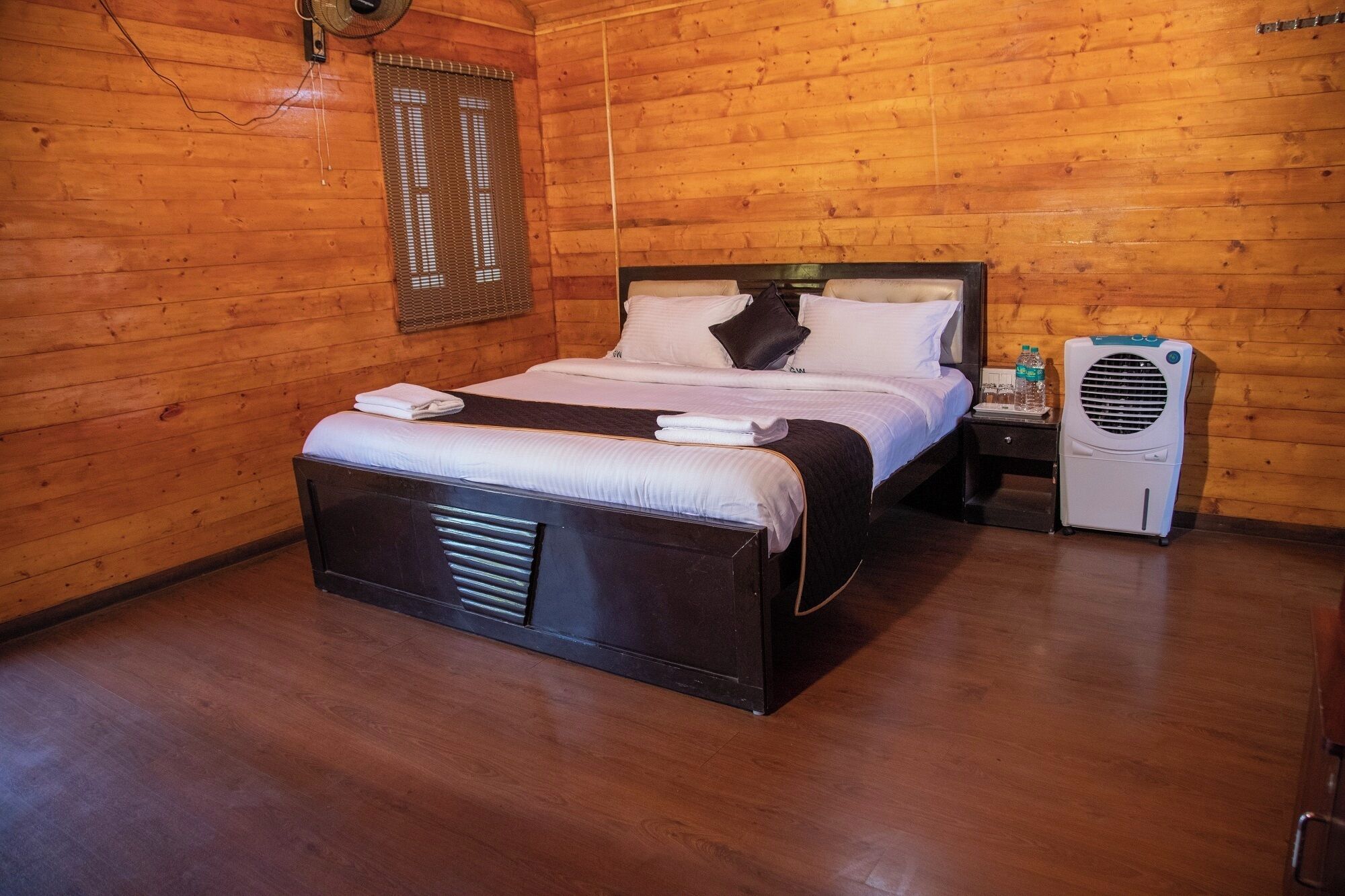 A wooden cabin room with a bed, a nightstand, a fan, and a window with blinds.