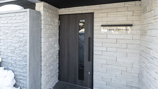 A modern entrance with a dark wooden door, white stone walls, and a sign above the door. - SERENADE Chitose MS103 (Chitose)