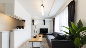 A modern living room with a sofa, a coffee table, a TV, and a large plant. - SERENADE Chitose MS103 (Chitose)