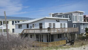 Een huis aan het strand met een balkon, een overdekte veranda en een zandstrand op de voorgrond.