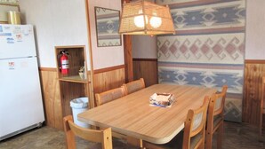Een houten eettafel met vier stoelen, een hanglamp, een brandblusser en een koelkast in een kamer met behang en houten lambrisering.