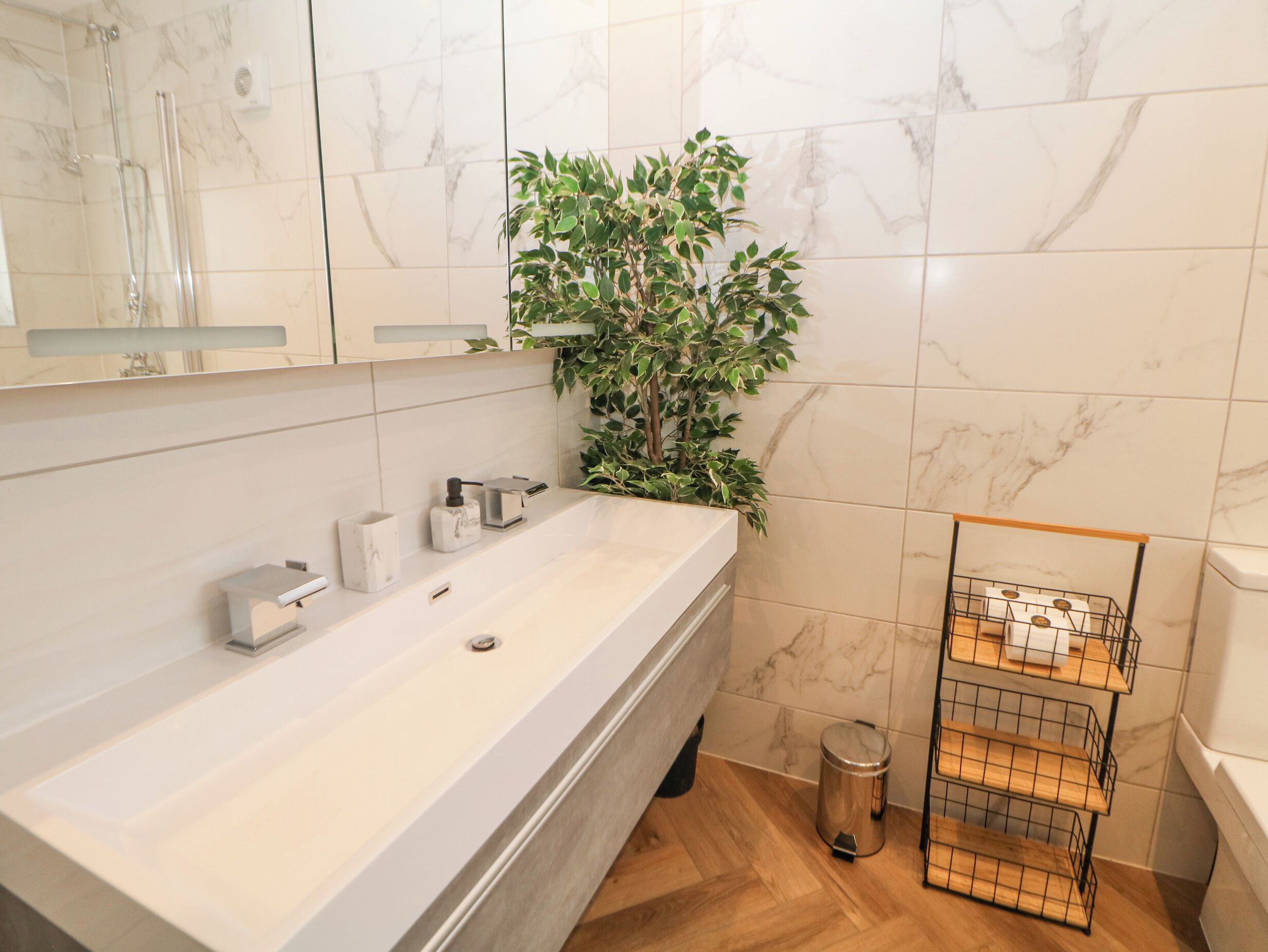 Un baño con pared de azulejos de mármol, un lavabo blanco, un espejo, una planta y un cubo de metal.