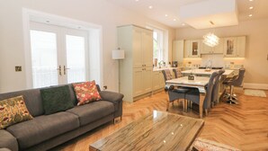 A modern living room with a gray sofa, a glass coffee table, and a dining area with a white table and bar stools. - Cavendish Suite (Buxton)