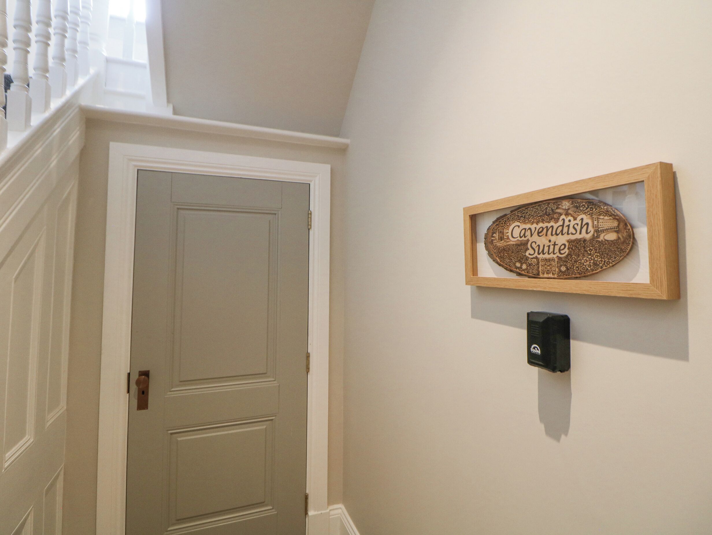 A hallway with a closed door, a framed sign reading "Cavendish Suite," and a wall-mounted device.