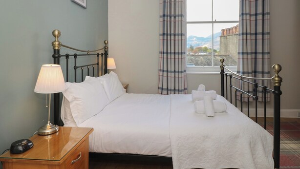 A neatly made bed with white linens, a bedside lamp, and a window with plaid curtains. - Baystones (Keswick)