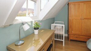 A room with a sloped ceiling, a wooden desk with a lamp, a crib, and a wooden wardrobe. - Baystones (Keswick)