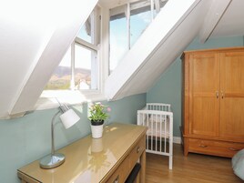 A room with a sloped ceiling, a wooden desk with a lamp, a crib, and a wooden wardrobe.