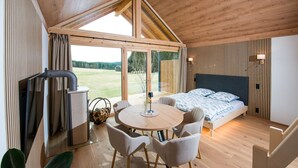 A modern bedroom with a wooden ceiling, a large bed with a blue and white patterned bedspread, a round wooden table with chairs, and a view of a grassy field through large windows. - Neu Gebautes Chalet im Bayerischen Wald (Hohenau)