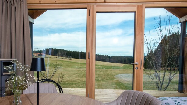 A room with a wooden table, a chair, a lamp, and a view of a grassy field through large glass doors. - Neu Gebautes Chalet im Bayerischen Wald (Hohenau)