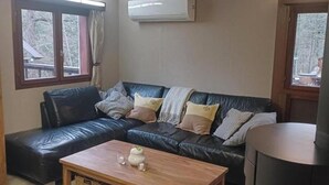 Una sala de estar con un sofá de cuero negro, una mesa de centro de madera y una pequeña mesa con velas. Hay una ventana con cortinas y un equipo de aire acondicionado en la pared.