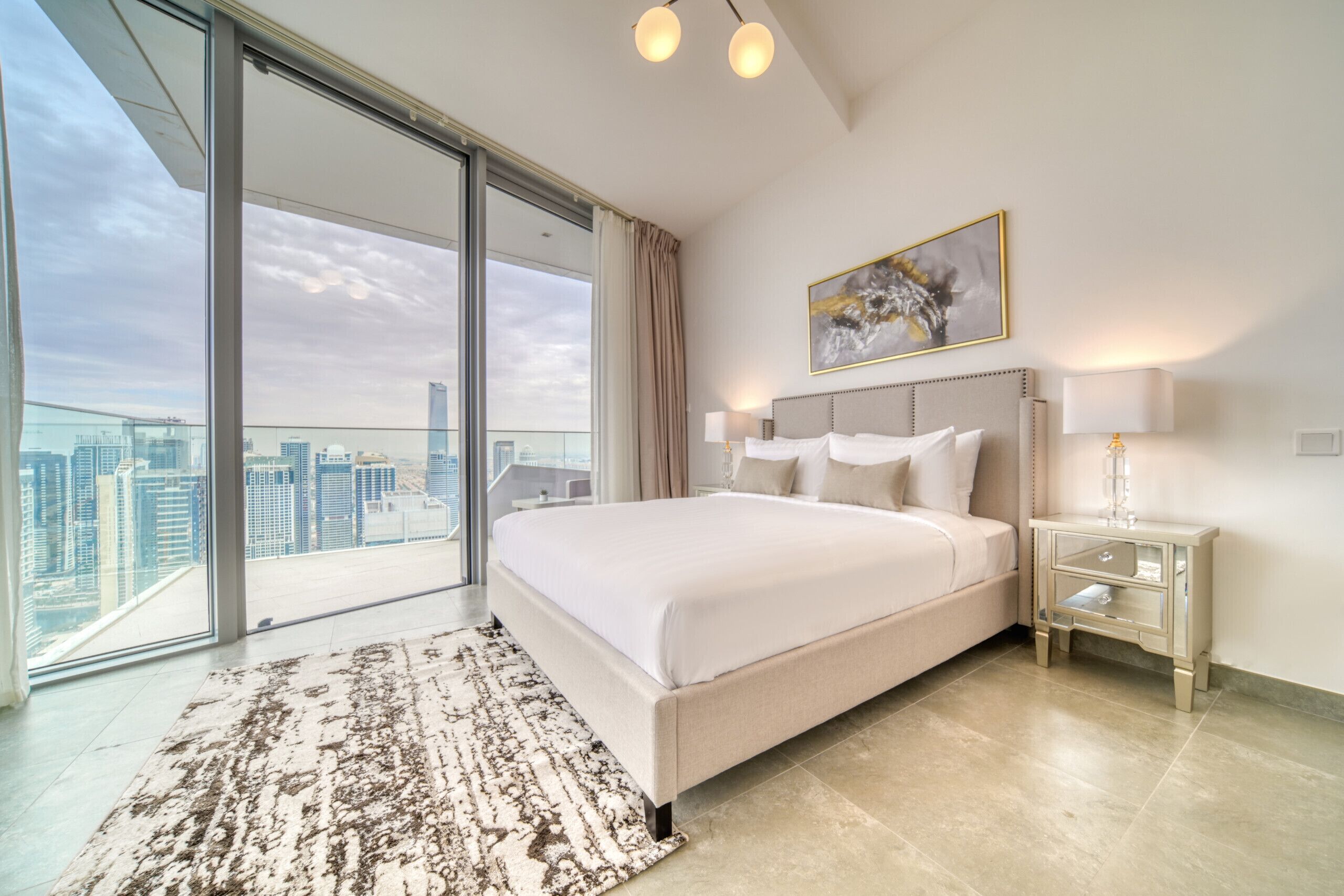 A modern hotel room with a large bed, a city view, and a glass door leading to an outdoor balcony.