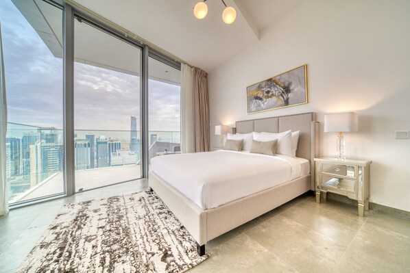 A modern hotel room with a large bed, a city view, and a glass door leading to an outdoor balcony.