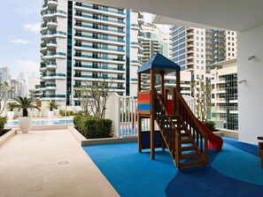 A children's play area with a slide and climbing structure, surrounded by a pool and potted plants.
