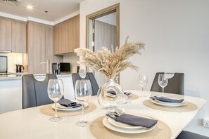 A modern dining area with a white table set for four, including wine glasses, plates, and cutlery. A decorative vase with pampas grass is on the table. The room has wooden cabinets and a kitchen in the background.