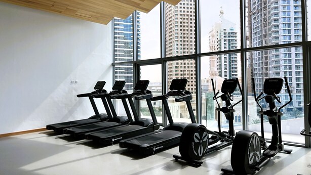 Un gimnasio con cintas de correr, una bicicleta estática y una máquina de remo, con amplias ventanas que ofrecen vistas a edificios altos.